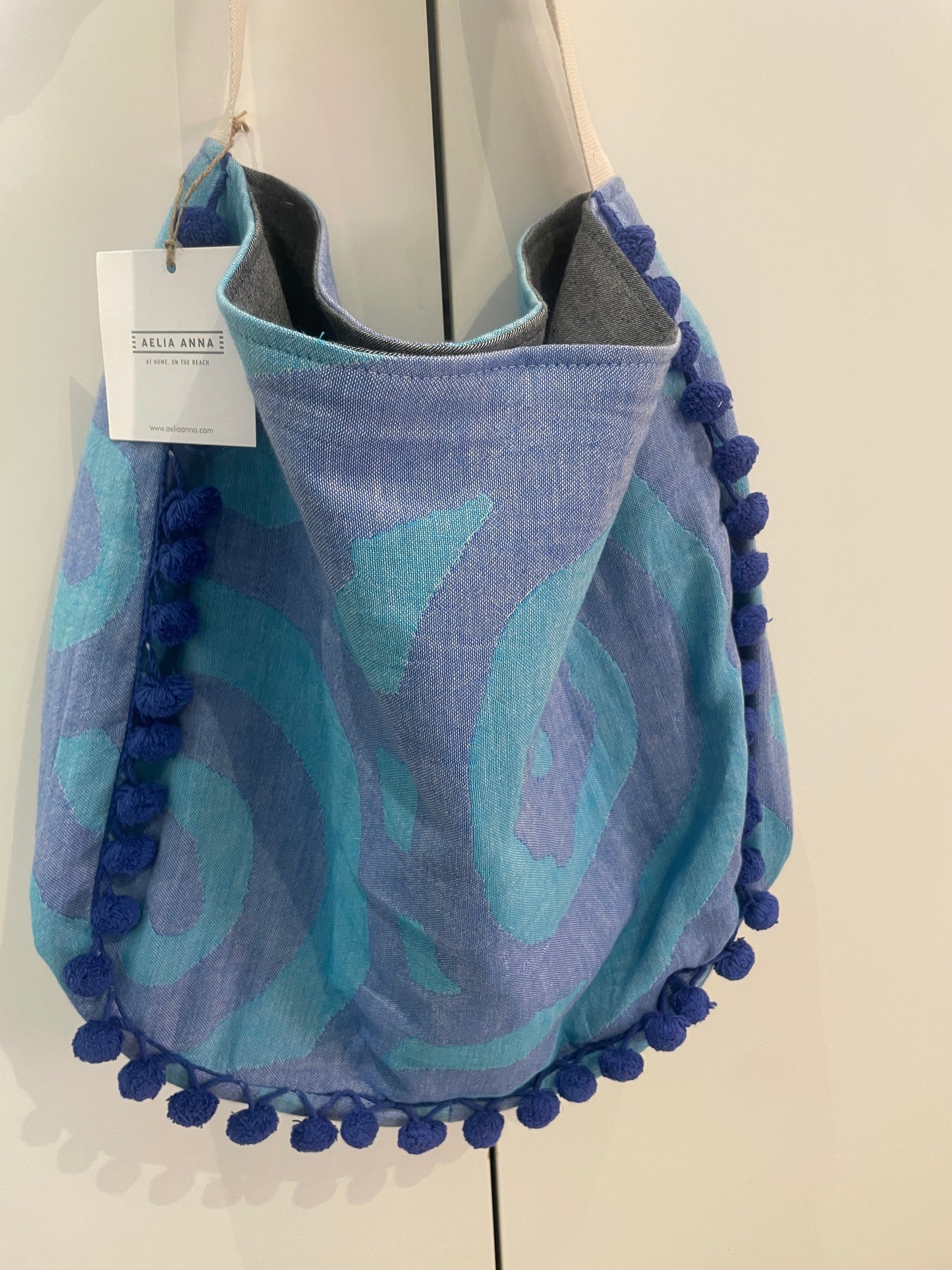 Borsa mare Aelia Anna blu e turchese con pompon decorativi e manici in tessuto, ideale per spiaggia, piscina e vacanze estive.