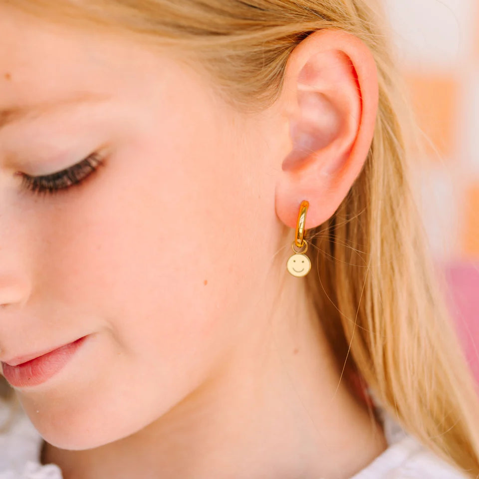 Bambina con i Yellow Smiley Charms in smalto giallo placcati oro 18K applicati agli Huggies Pierced Co, ipoallergenici e sicuri per orecchie sensibili.